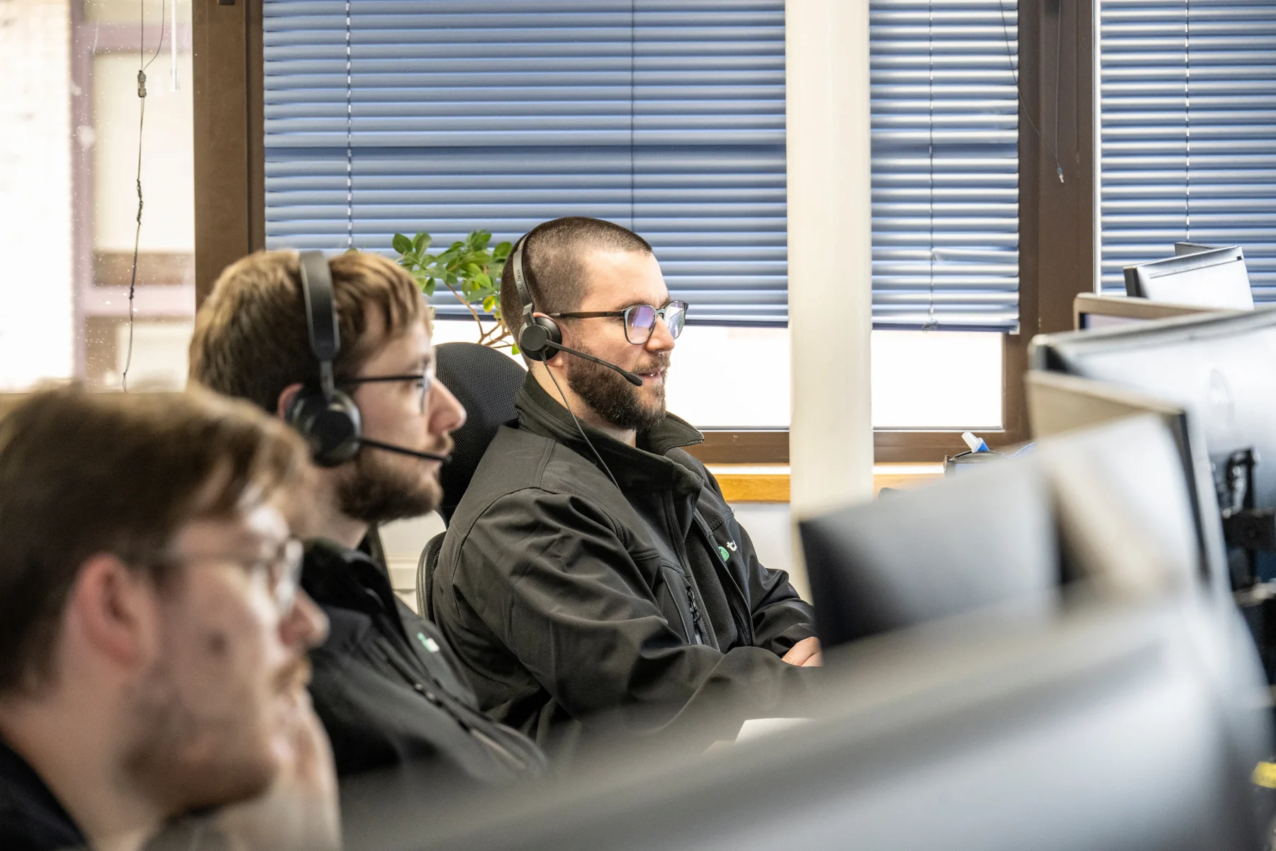 Three office workers wearing headsets sit at computer workstations in a modern office, assisting customers while looking at their screens.
