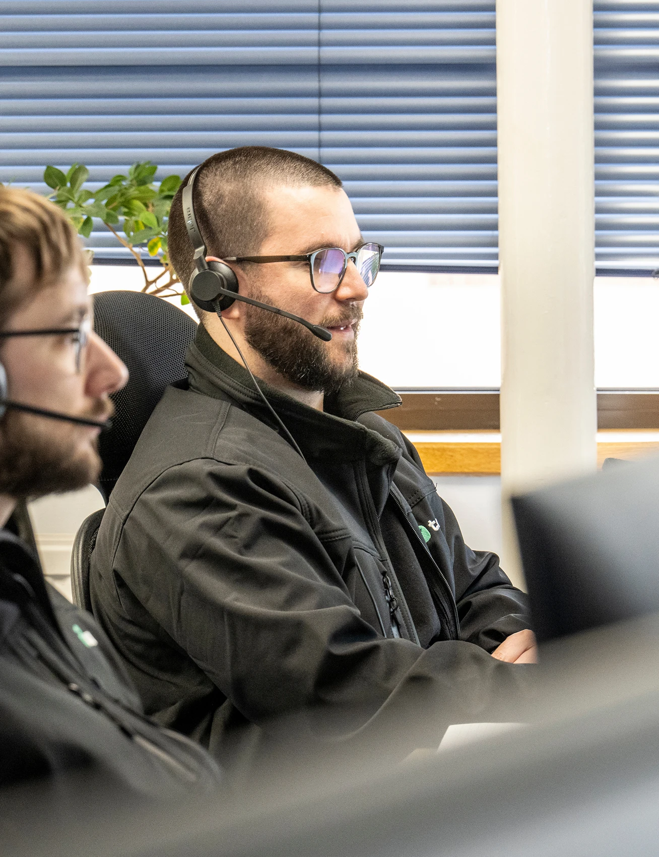 Three office workers wearing headsets sit at computer workstations in a modern office, assisting customers while looking at their screens.