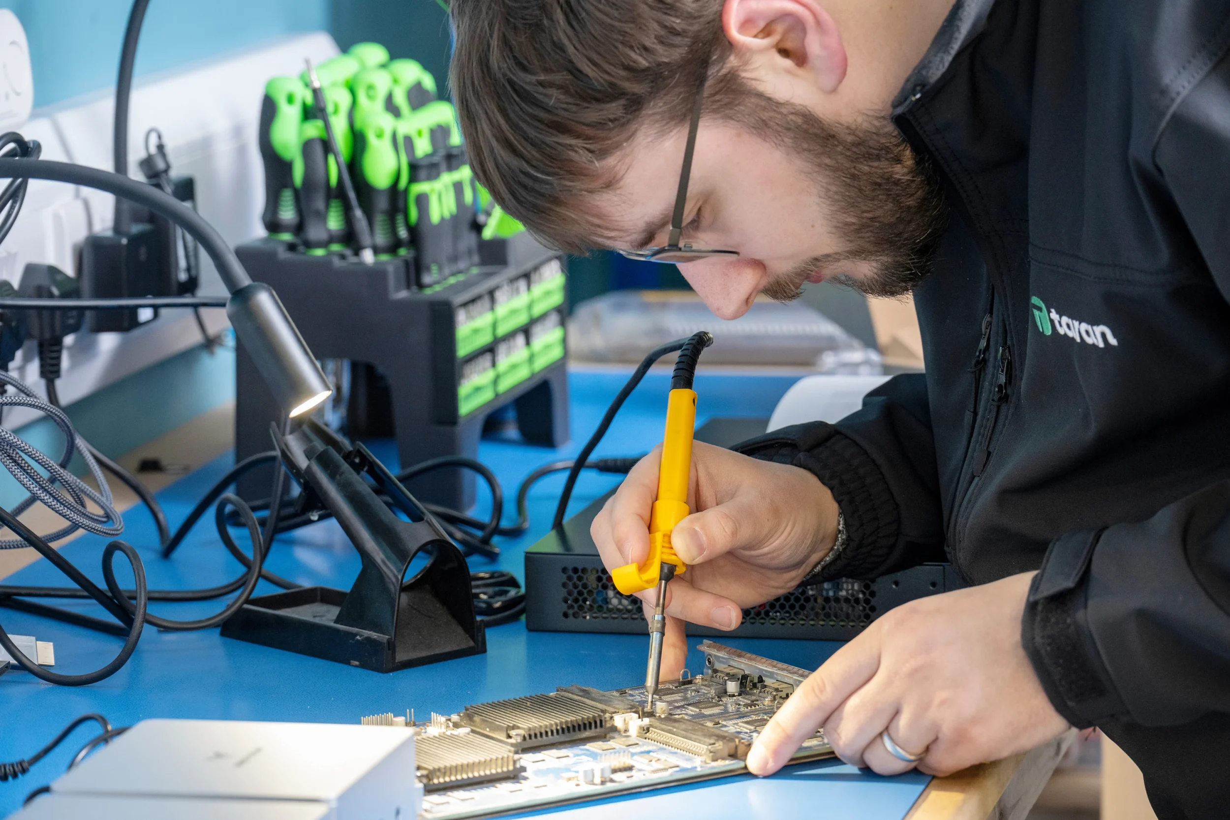 Taran IT technician repairing computer hardware using a soldering iron at a workbench.
